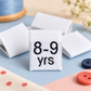 White 8–9 years kids’ woven size label displayed on a pastel blue and pink sewing background