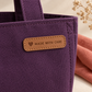 Tan custom leather label reading Made with Care, stitched onto a purple canvas tote bag