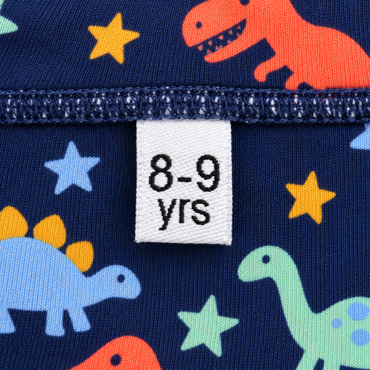 White 8–9 years kids’ woven clothing size label sewn into a navy dinosaur-print garment